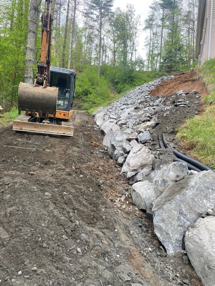 An excavator grading a roadbed beside a freshly built riprap stone wall on a wooded slope