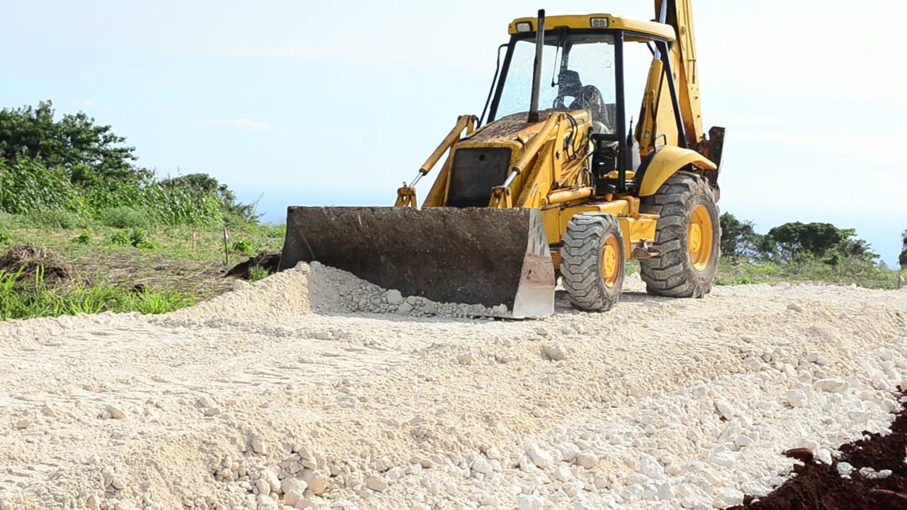 A backhoe loader grading a gravel pad on a hilltop under a blue sky